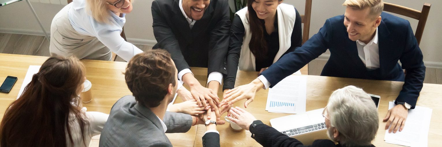 Young and old multiracial business people join pile of stacked hands together at group meeting conference table promising help support, concept of successful teamwork, team building unity, top view